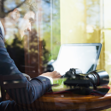 Young man photographer working at cafe, using laptop. His dslr camera at the table. Image through the windowの写真素材