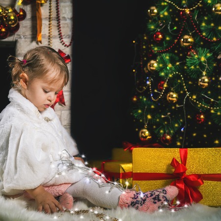 Cute toddler girl playing near the fireplace and decorated Christmas tree. New Year 2017. Dressed in fashion fur coatの写真素材