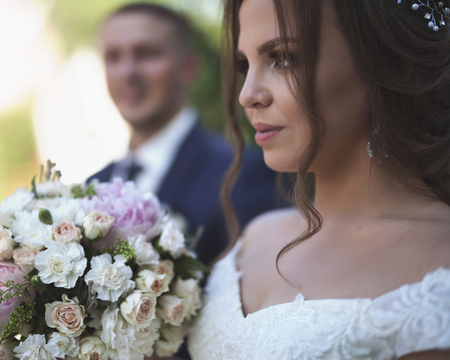 Portrait of Bride holds wedding bouquet and groom at background.