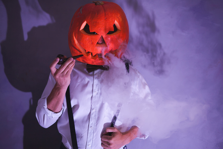 Handsome Man with carved pumpkin on his head for Halloween party smokes smoking pipeの写真素材