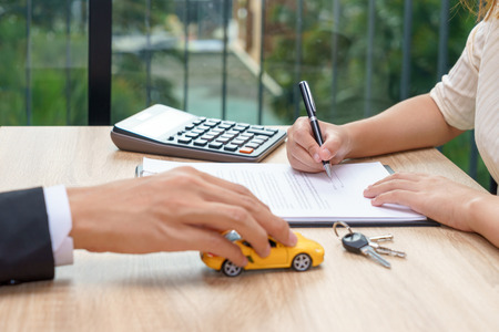 Woman signing car loan agreement contract with car key and calculator on wooden desk