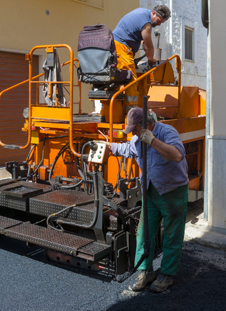 workers on a paver machine control the drafting of a new layer of asphalt for maintenance of city streetsの写真素材