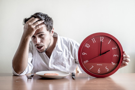 young stylish man with white shirt holding red clock behind a tableの写真素材