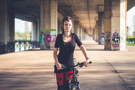young beautiful punk dark girl riding bike in urban landscapeの写真素材