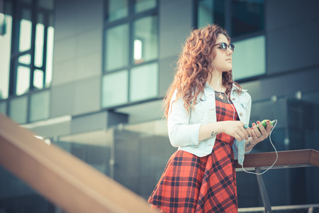 young beautiful hipster woman with red curly hair listening music in the cityの写真素材