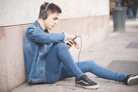 young handsome alternative dark model man in town listening to music with headphones and smartphone connected wirelessの写真素材