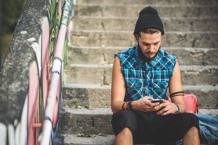 young handsome bearded hipster man using smartphone and listening music with earphones in the cityの写真素材