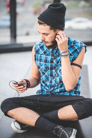 young handsome bearded hipster man using smartphone and listening music with earphones in the cityの写真素材