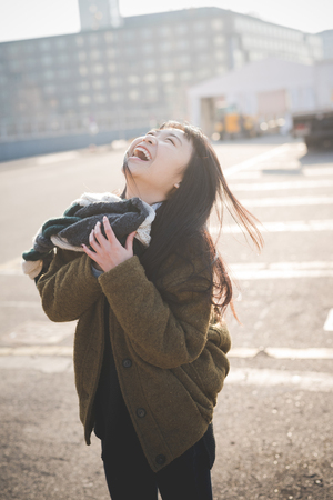 young beautiful asian hipster woman in the city - living town during sunset in backlit - smiling happy having fun with scarfの写真素材