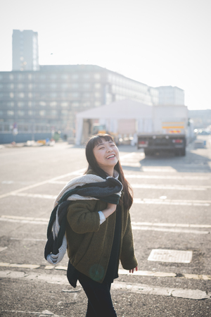 young beautiful asian hipster woman in the city - living town during sunset in backlit - smiling happy having fun with scarfの写真素材