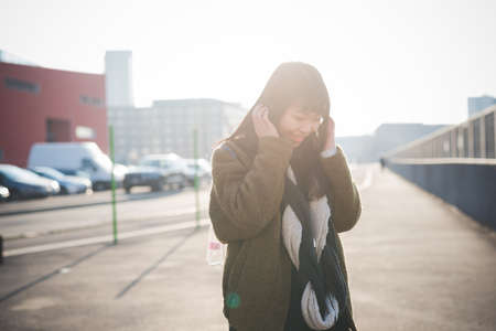 young beautiful asian hipster woman in the city - living town during sunset in backlit - listening music with headphonesの写真素材