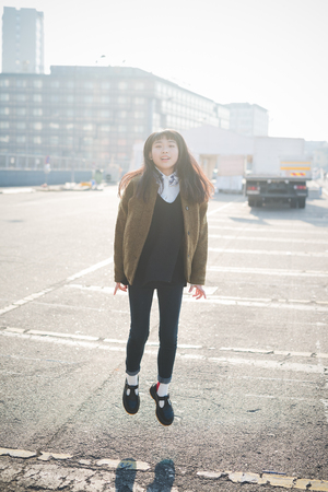 young beautiful asian hipster woman in the city - living town during sunset in backlit jumping and dancingの写真素材