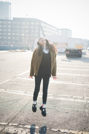 young beautiful asian hipster woman in the city - living town during sunset in backlit jumping and dancingの写真素材