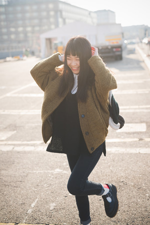 young beautiful asian hipster woman in the city - living town during sunset in backlit - smiling happy having fun with scarfの写真素材