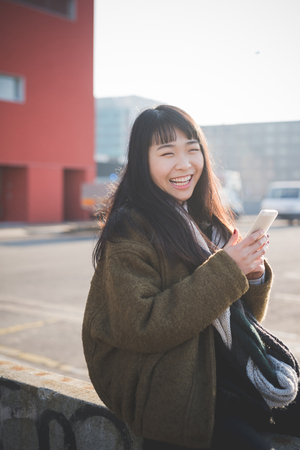 young beautiful asian hipster woman in the city - living town during sunset in backlit - using device smartphone connected onlineの写真素材