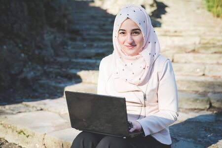 young beautiful muslim woman at the park in spring using notebook connected online workingの写真素材