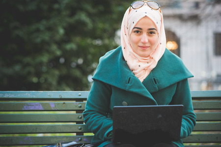 young beautiful muslim woman at the park in spring using notebook connected online working businessの写真素材