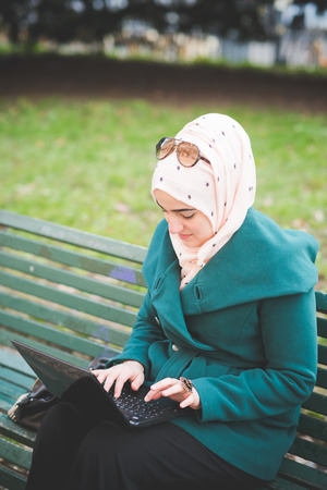 young beautiful muslim woman at the park in spring using notebook connected online working businessの写真素材