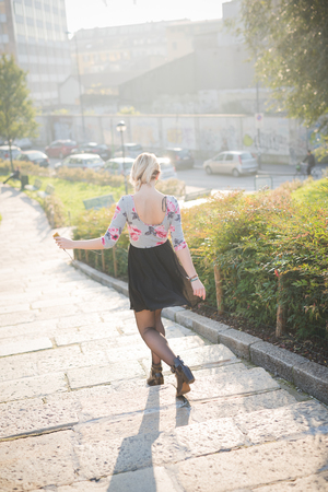 Young and beautiful caucasian blonde girl walking through the city at dusk wearing a floral dress with a black skirt.の写真素材