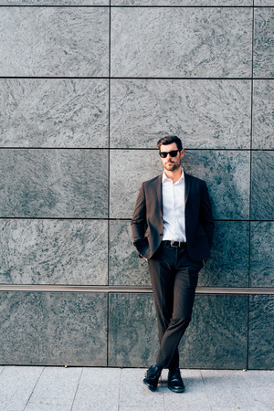 Young handsome modern businessman posing leaning against a wall, looking in camera, wearing a coat - business, success conceptの写真素材