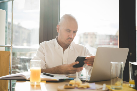 Half length of young handsome caucasian bald business man sitting in a bar using a laptop leaning on his knees, looking downward the screen, pensive - working, thoughtful, busy conceptの写真素材