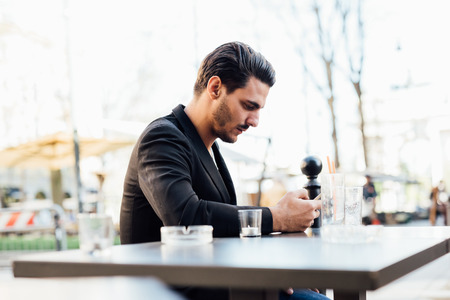 Young handsome italian boy seated on a bar in the city center using a smartphone looking downward the screen - social network, communication, technology conceptの写真素材