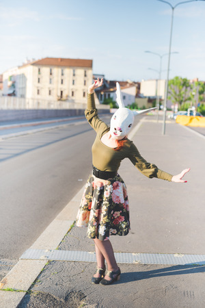 young handsome caucasian redhead woman with rabbit mask, dancing in the street of the city, holding a strip of her floral skirt - absurd, strange, dreamlike conceptの写真素材