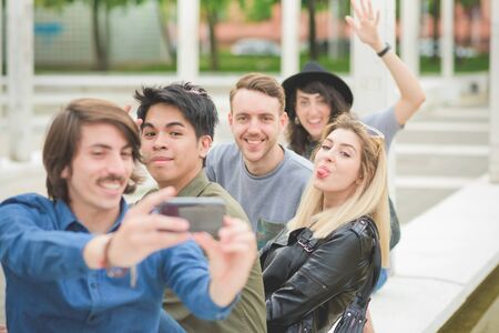 Group of young multiethnic friends sitting on a small wall, talking to each other, using a smartphone, taking a selfie , having fun - friendship, relaxing conceptの写真素材