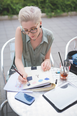 Half length of a young handsome caucasian blonde italian designer sitting in a bar, drawing on her sketchbook, using tablet and smartphone too - technology, creativity, working conceptの写真素材