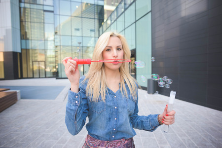 Young handsome blonde caucasian girl having fun playing with soap bubble in the city wearing jeans...