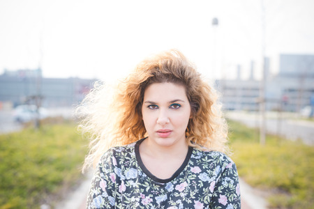 Portrait of young curly blonde hair caucasian girl posing in the street of the city, looking in camera, serious - pensive, youthful, thinking future conceptの写真素材