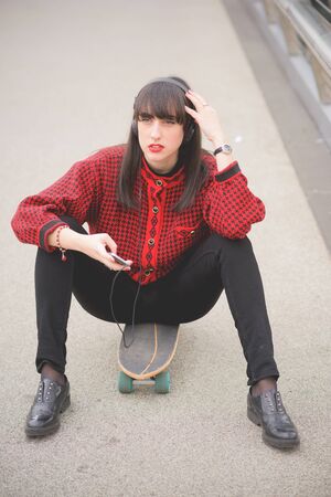 Young beautiful caucasian woman seated on a skate using a smartphone listening to music overlooking - technology, social network, communication conceptの写真素材