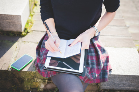 close up on woman hands using tablet, smartphone, and writing on notebook with a pen leaning on her knees - technology, multitasking, working conceptの写真素材