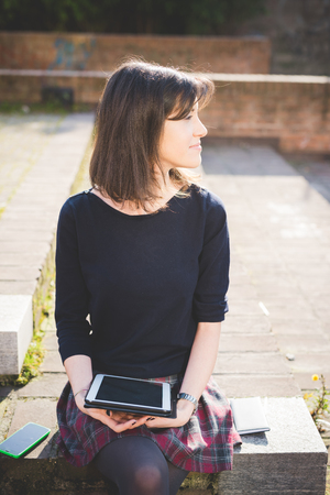 young pretty brown hair woman sitting on a sidewalk, in town, using a tablet, overlooking left - technology, multitasking, social network conceptの写真素材