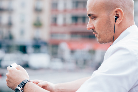 Half length of young handsome caucasian bald business man listening music with earphones, holding a smartphone, looking downward the screen - music, relaxing conceptの写真素材