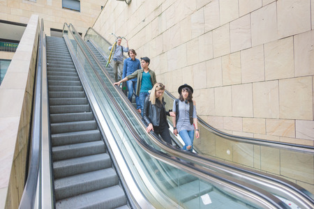 Group of young multiethnic friends going downwstairs on a escalator holding skateboards and bicycle,...