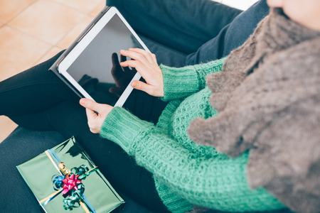 Close up on the hand of young handsome woman tapping on the screen of a tablet sitting on the sofa, gift place on her side- technology, communication, social network conceptの写真素材