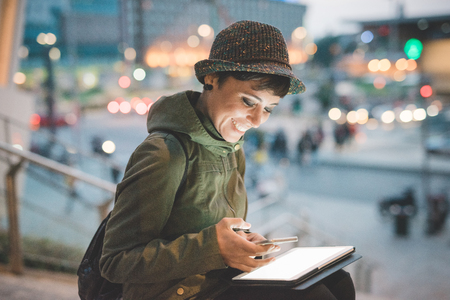 Young handsome caucasian brown straight hair woman sitting on a staircase at nightfall, tablet on her knee, smartphone handheld, face enlightened by the light of the screen, smiling -の写真素材