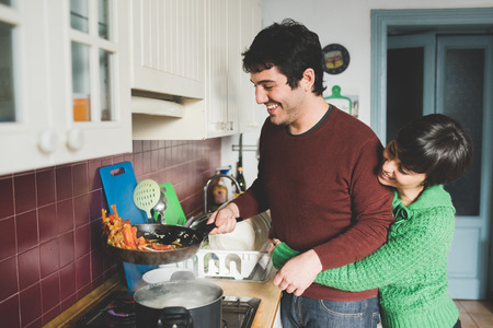 Half length of young handsome caucasian man and woman couple cooking together, she is hugging him form back and he is tossing a pan with vegetables - cooking, food, love conceptの写真素材