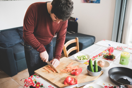 Young handsome caucasian brown hair man cutting some carrots with knife on a cutting board, surrounding by bowl, vegetables, pan and knife - healthy, veggie, food conceptの写真素材