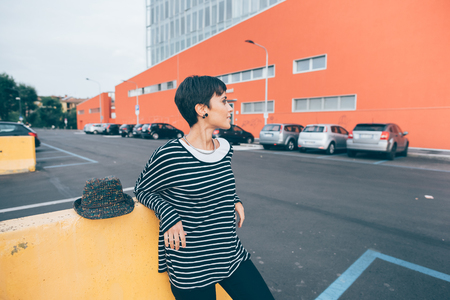 Knee figure of young handsome caucasian brown straight hair woman leaning against a small wall in the city overlooking left smiling - serene, relax, carefree conceptの写真素材