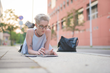 young handsome caucasian blonde italian designer lying down on a sidewalk, usin a tablet, looking downward and tapping the screen - technology, social network, communication conceptの写真素材