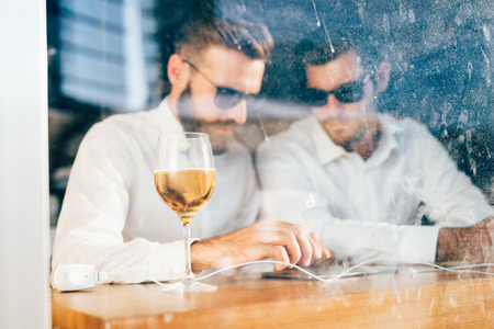 Two young bearded caucasian modern business man sitting in a bar, using tablet looking downward,...