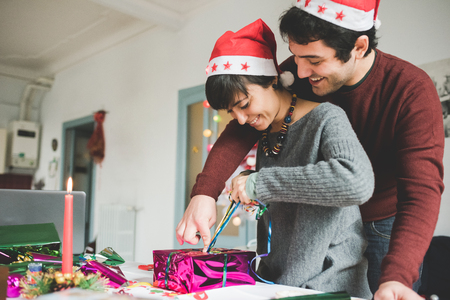 Half length profile of young handsome caucasian man and woman couple wrapping christmas present with wrapping paper and ribbon he is hugging her from back, smiling - christmas, couple, holiday conceptの写真素材