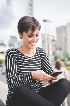 young handsome caucasian brown straight hair woman sitting on a small wall, leg crossed, holding a smartphone, looking down the screen - technology, social network, communication conceptの写真素材