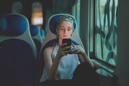 half length of a young handsome caucasian blonde italian designer sitting in a train, holding a smartphone, looking down the screen, smiling - technology, social network, communication conceptの写真素材