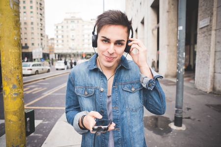 Half length of young handsome alternative dark model man in town listening to music with headphones and smartphone, looking in camera smiling - music, technology, relaxing conceptの写真素材