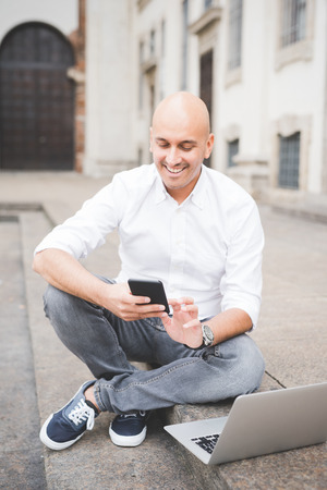 Young handsome caucasian business man sitting on a staircase using smartphone handhold and a laptop - working, multitasking, busy conceptの写真素材