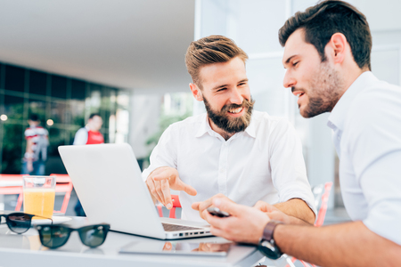 Two young bearded caucasian modern business man sitting in a bar, using laptop, looking each other, smiling and chatting - business, work, technology conceptの写真素材