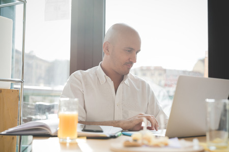 Half length of young handsome caucasian bald business man sitting in a bar using a laptop looking downward and tapping the screen, smiling - working, happy, busy conceptの写真素材